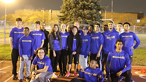 A group of eighteen individuals from Kilomaîtres LaSalle, wearing blue shirts, pose together outdoors at night on a track field. Some are standing while others kneel. Trees and a fence create a picturesque backdrop.