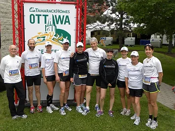 A group of nine people in running gear stand together for a photo in front of a sign that reads "Tamarack Homes Ottawa" outdoors, ready for their marche rapide.