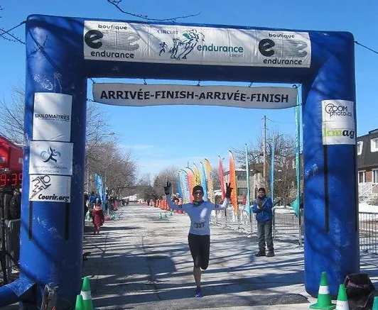 A runner crosses the finish line at a Course Populaire race, arms raised in celebration, with a blue inflatable arch overhead displaying "Arrivée-Finish-Arrivée-Finish".