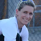 Francisca Russli, with her blonde hair and sunglasses, smiles while sitting in front of a chain-link fence, dressed in a white top.