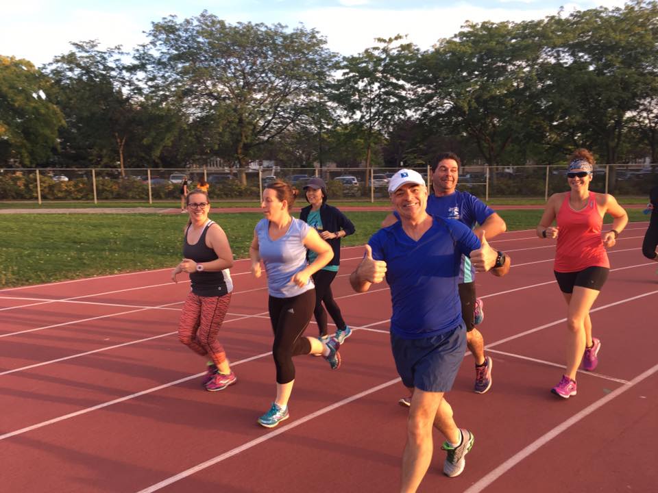 A group of people from Le Club jogs on a running track in an outdoor setting with trees in the background. One person in the foreground is giving a thumbs-up sign while smiling.