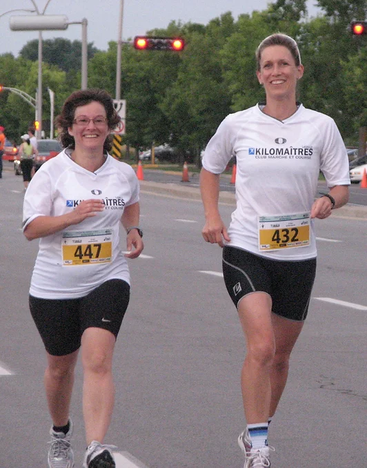 Two women, wearing white "Kilomaitres" shirts and numbered bibs, are engaging in a marche rapide during an outdoor race event. They are smiling and jogging on a road with other participants in the background.