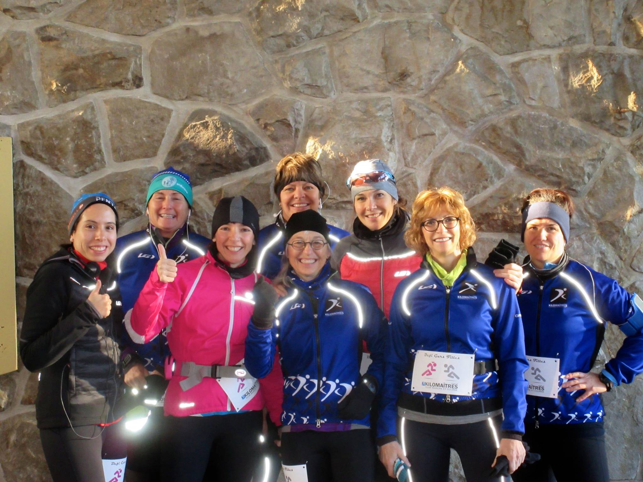 A group of nine runners in athletic gear, some wearing numbered bibs, smiles and poses for a photo against a stone wall, ready for their marche rapide.