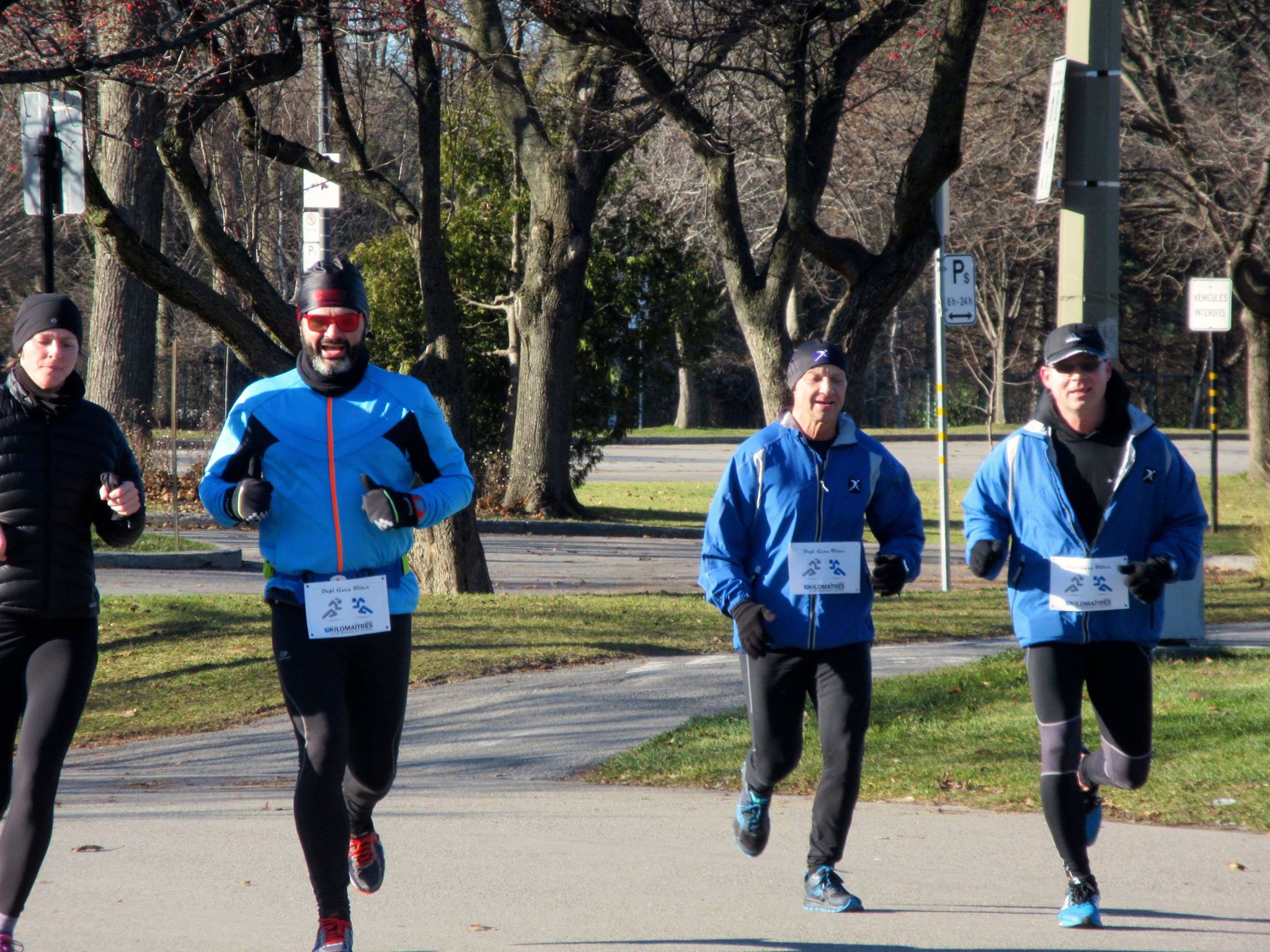 Four runners are engaging in a marche rapide on a park path during a race, wearing race bibs and cool weather gear.