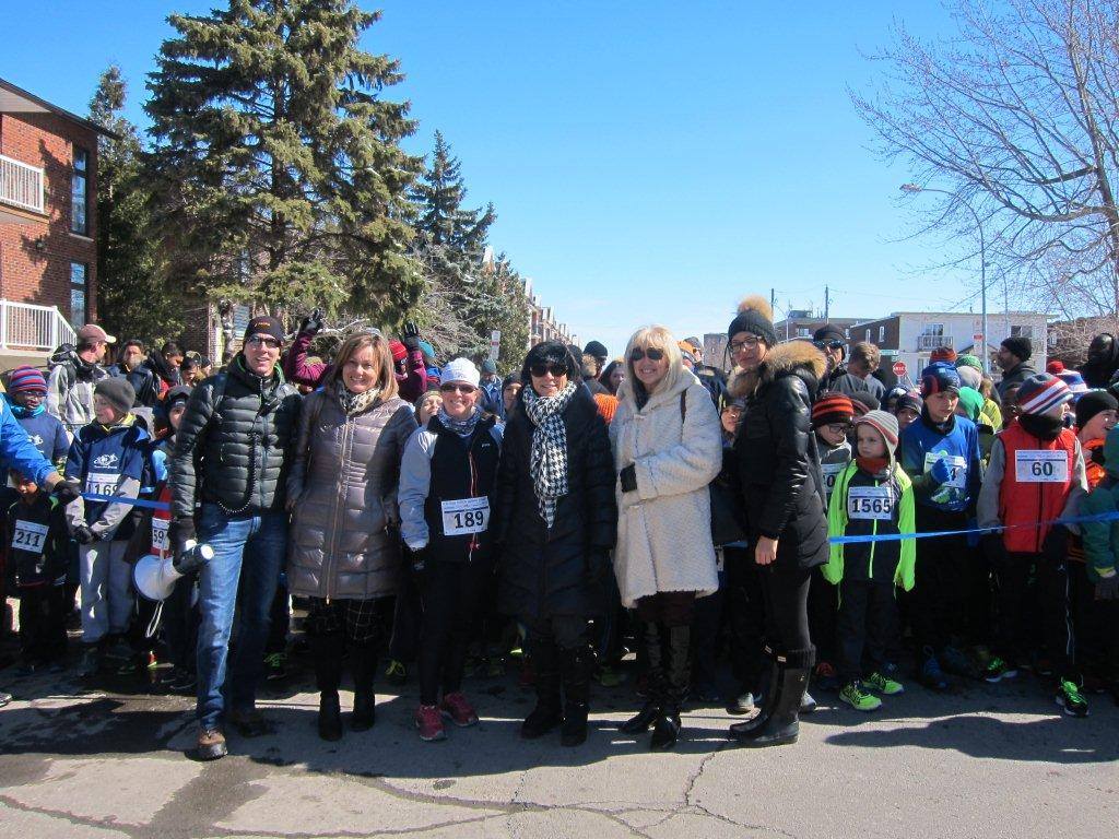 A group of adults and children, many wearing winter clothing and race numbers, stand at the starting line of a marche rapide event on a bright day. Trees and buildings are visible in the background.