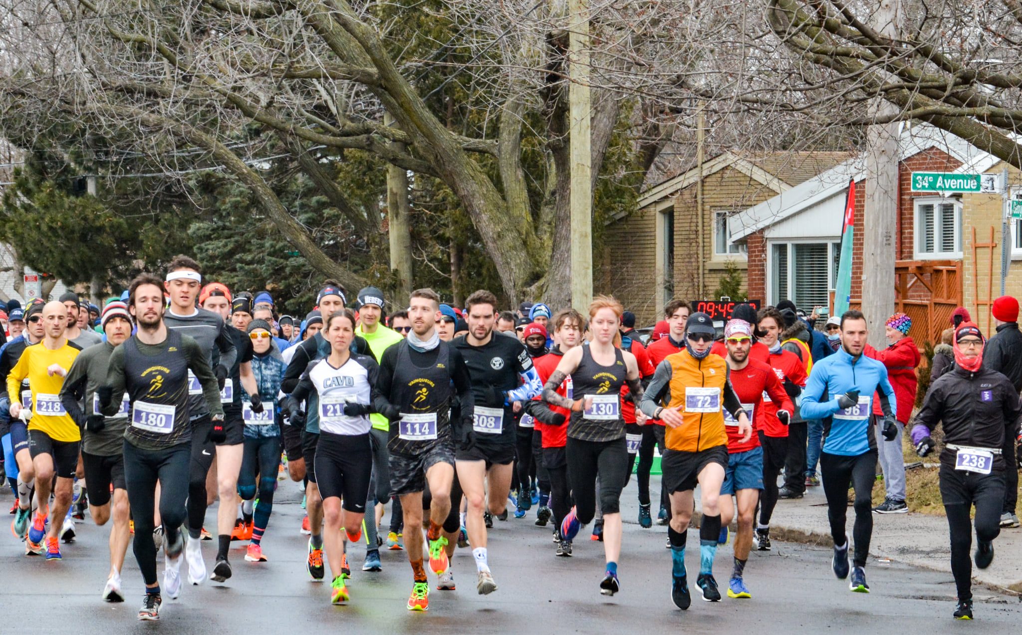 A large group of runners participating in a street race on a cold day, with some spectators in the background and bare trees lining the street, resembles a scene from a marche rapide event.