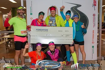 A group of smiling people in brightly colored athletic attire from Le Club hold a large ceremonial check, posing for a photo at an event. Logos for a foundation and sponsors are visible in the background.