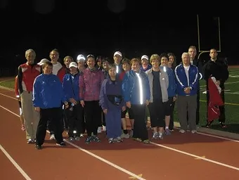 A group of people standing together on an outdoor track at night, dressed in athletic wear, appears ready for a vigorous session of marche rapide.