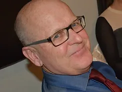 A bald man with glasses and a grey short beard, resembling Mike Strange, is smiling slightly while looking into the camera. He is wearing a blue shirt and a red tie.