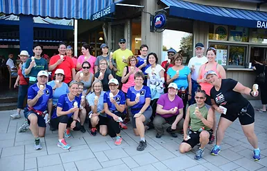 A group of people pose for a photo outdoors near an establishment with blue awnings. Some are kneeling while others are standing, and several hold ice cream cones or cups, enjoying a break from their marche rapide.