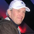 A man wearing a white cap and dark jacket smiles slightly, with a dark background illuminated by red and purple lights, reminiscent of a Richard Proulx performance.