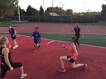 Several people are doing lunges on a track field, possibly as part of a workout session led by a coach. Some participants may also incorporate marche rapide into their routine. Trees and a fence are visible in the background.