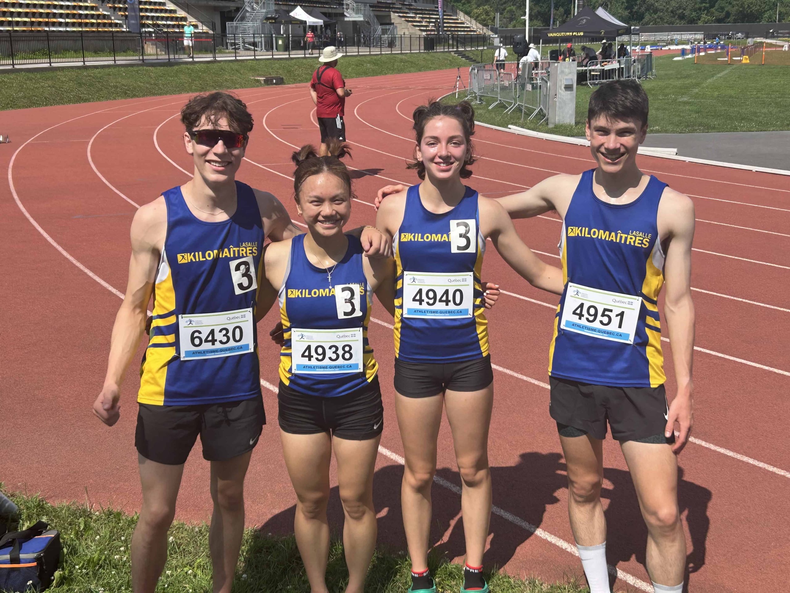 Four athletes from the club d’athlétisme, in matching running attire with race numbers, pose on a track. The athletes' uniforms are blue with yellow side panels, and the photo is taken on a sunny day at a race event.