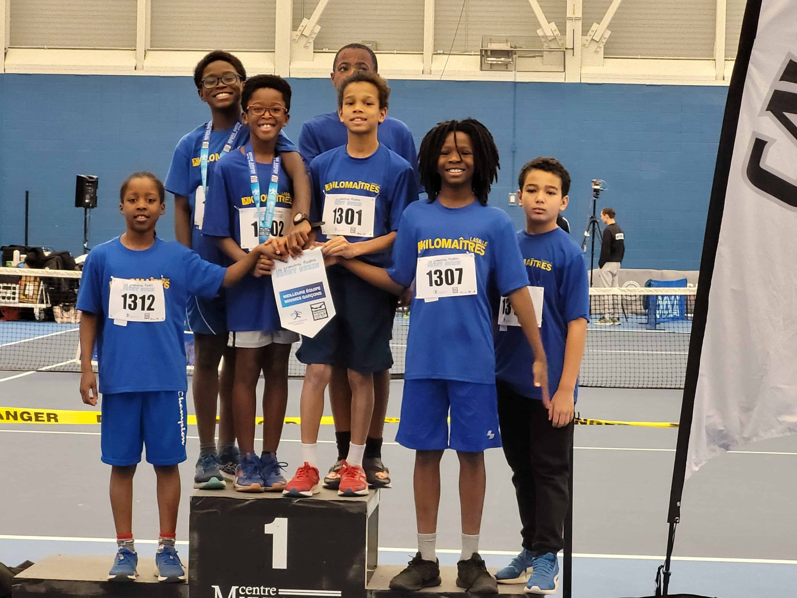 A group of young athletes in blue shirts stand on a podium, representing their club d’athlétisme. The center athlete proudly holds a small banner. The setting appears to be an indoor sports facility.