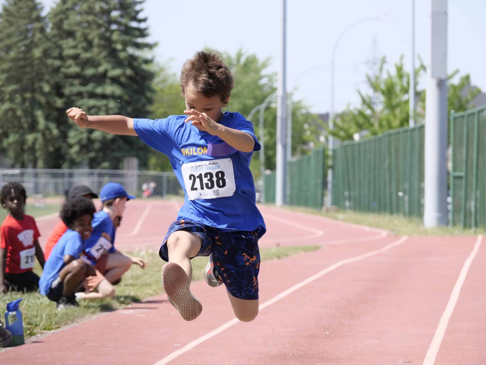 A young child wearing a blue shirt and shorts jumps in the air while competing in a race on a track, representing their club d’athlétisme. Spectators can be seen sitting on the grass in the background.