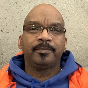 A man with glasses, a goatee, and a bald head—resembling Colin Keith—is wearing a blue hoodie and an orange jacket, standing in front of a textured, beige wall, Coaches