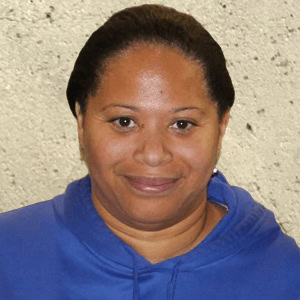 Ingrid, a woman with dark hair and a blue hoodie, smiles slightly in front of a textured light-colored wall, Coaches