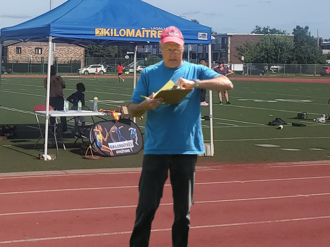 A man in a blue shirt and red cap holds a clipboard on a track field. In the background, a blue tent labeled "Kilomaitres" stands next to a table with items, representing the nearby club d’athlétisme.