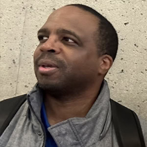 A man with short hair and a backpack, wearing a gray jacket, looks to his right against a speckled gray wall. Todd Bertrand, Coaches