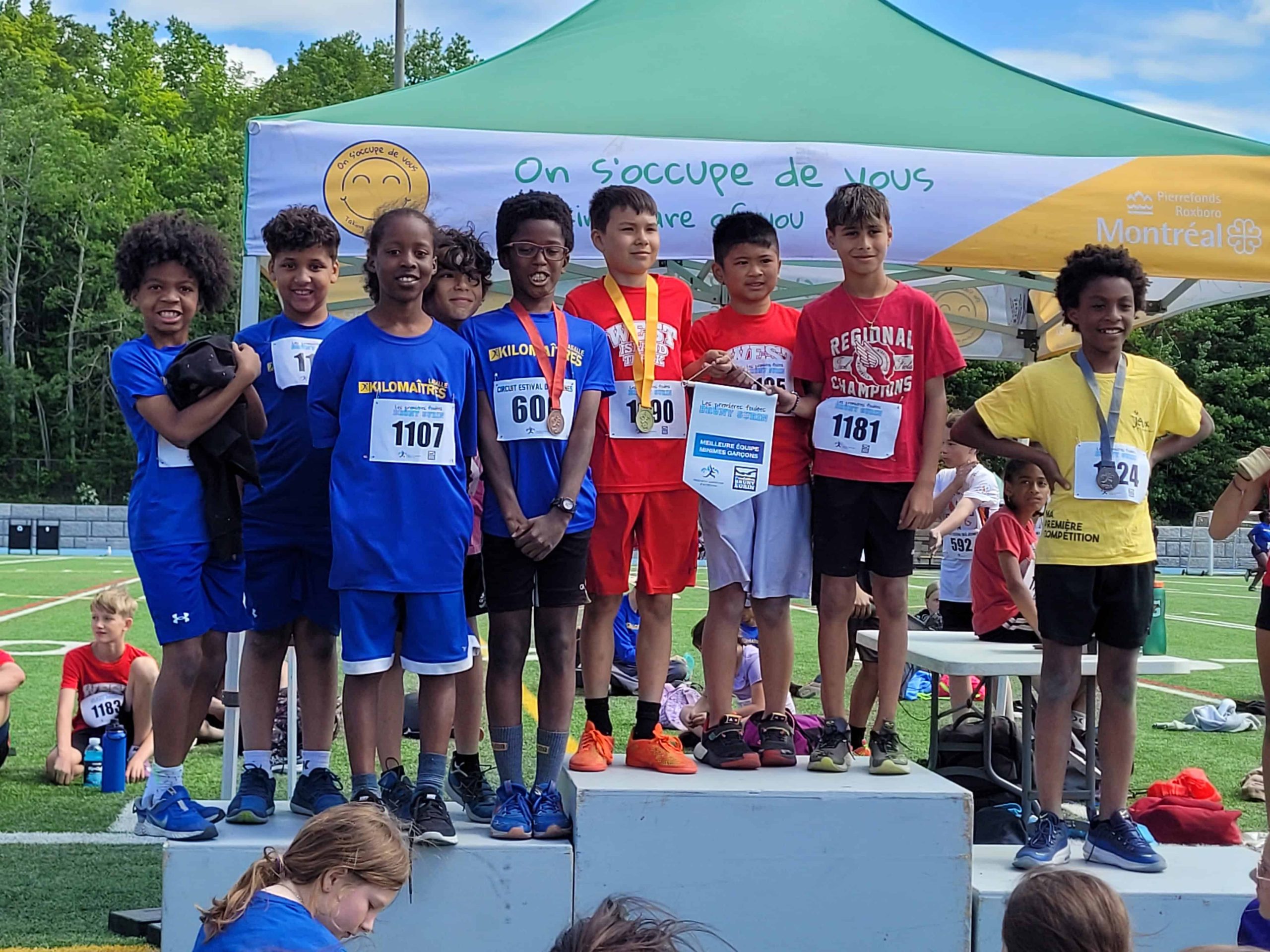 A group of young boys from Kilomaîtres LaSalle stand proudly on a podium, posing for a photo after a sporting event. Some are holding medals and certificates. A tent and several people are visible in the background.