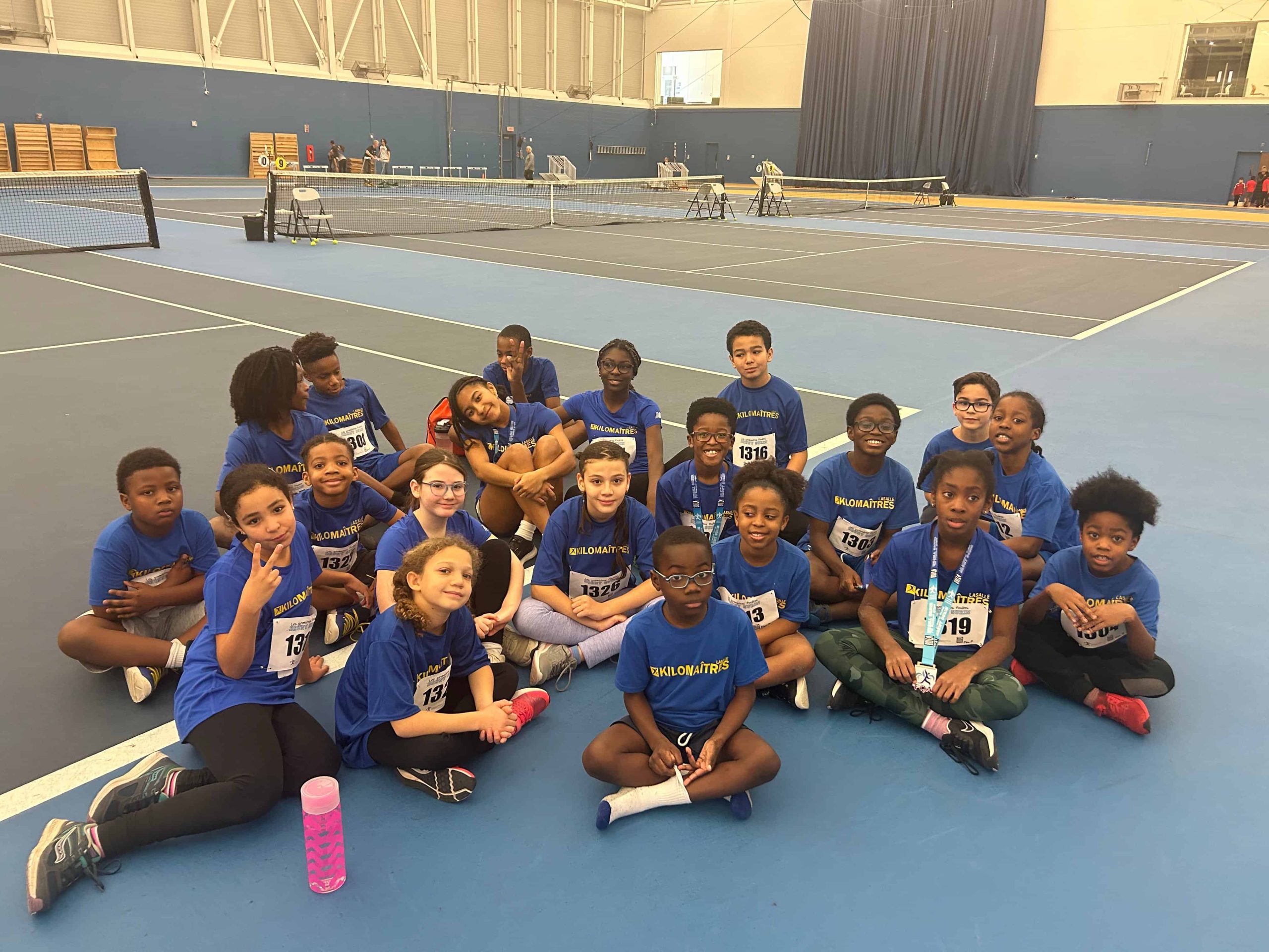 A diverse group of kids in blue shirts, some wearing numbered bibs, are sitting on the floor of the Kilomaîtres LaSalle indoor sports facility, possibly having finished or preparing for an activity.