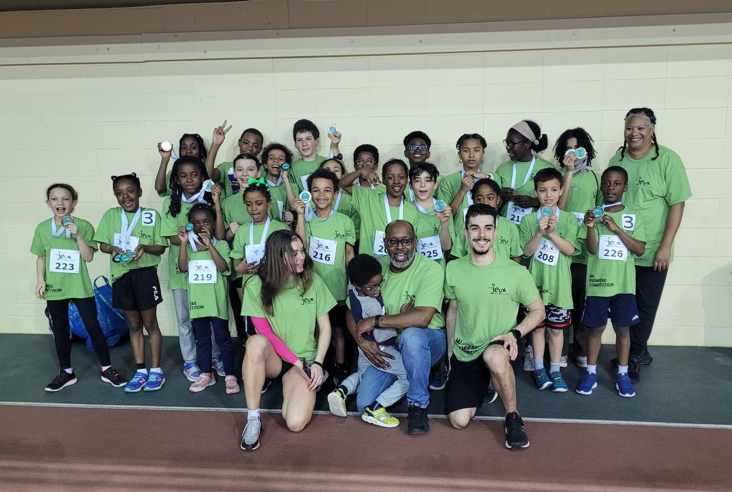 A group of children and adults in green shirts, representing Kilomaîtres LaSalle, posing with medals after a sports event in an indoor athletic facility.