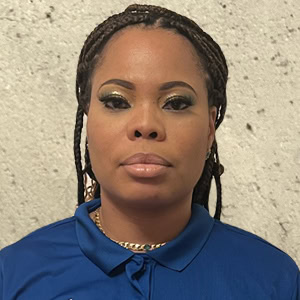 A person with braided hair, wearing a blue collared shirt, stands against a textured, light-colored wall, resembling the serene elegance often depicted in Lenore's timeless portraits, Coaches