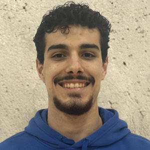 A man named Tristan, with short, curly hair and a beard, smiles while wearing a blue hoodie in front of a beige textured wall, Coaches