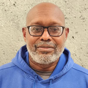 A man named Patrick, sporting a bald head, glasses, and a gray beard, is wearing a blue hoodie and standing against a concrete wall, Coaches